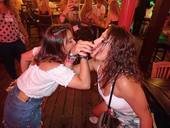 Two women taking a shot together at a crowded outdoor bar on a wooden deck, friends enjoying a lively nighttime party and nightlife scene.