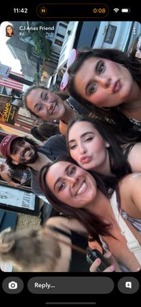 Group selfie of five young adults on a lively downtown street at dusk, smiling and making playful faces with storefronts, parked cars and an American flag in the background.