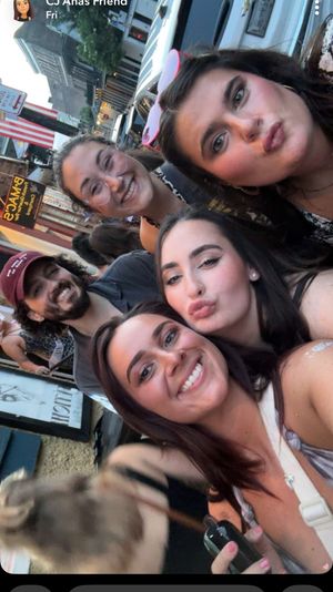 Group selfie of five young adults on a lively downtown street at dusk, smiling and making playful faces with storefronts, parked cars and an American flag in the background.