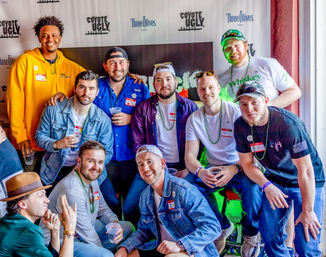 Group of smiling men posing at a lively indoor bar event, wearing casual clothes, bead necklaces and name tags, holding drinks in front of a step-and-repeat backdrop.