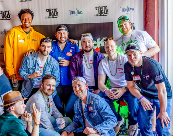 Group of smiling men posing at a lively indoor bar event, wearing casual clothes, bead necklaces and name tags, holding drinks in front of a step-and-repeat backdrop.