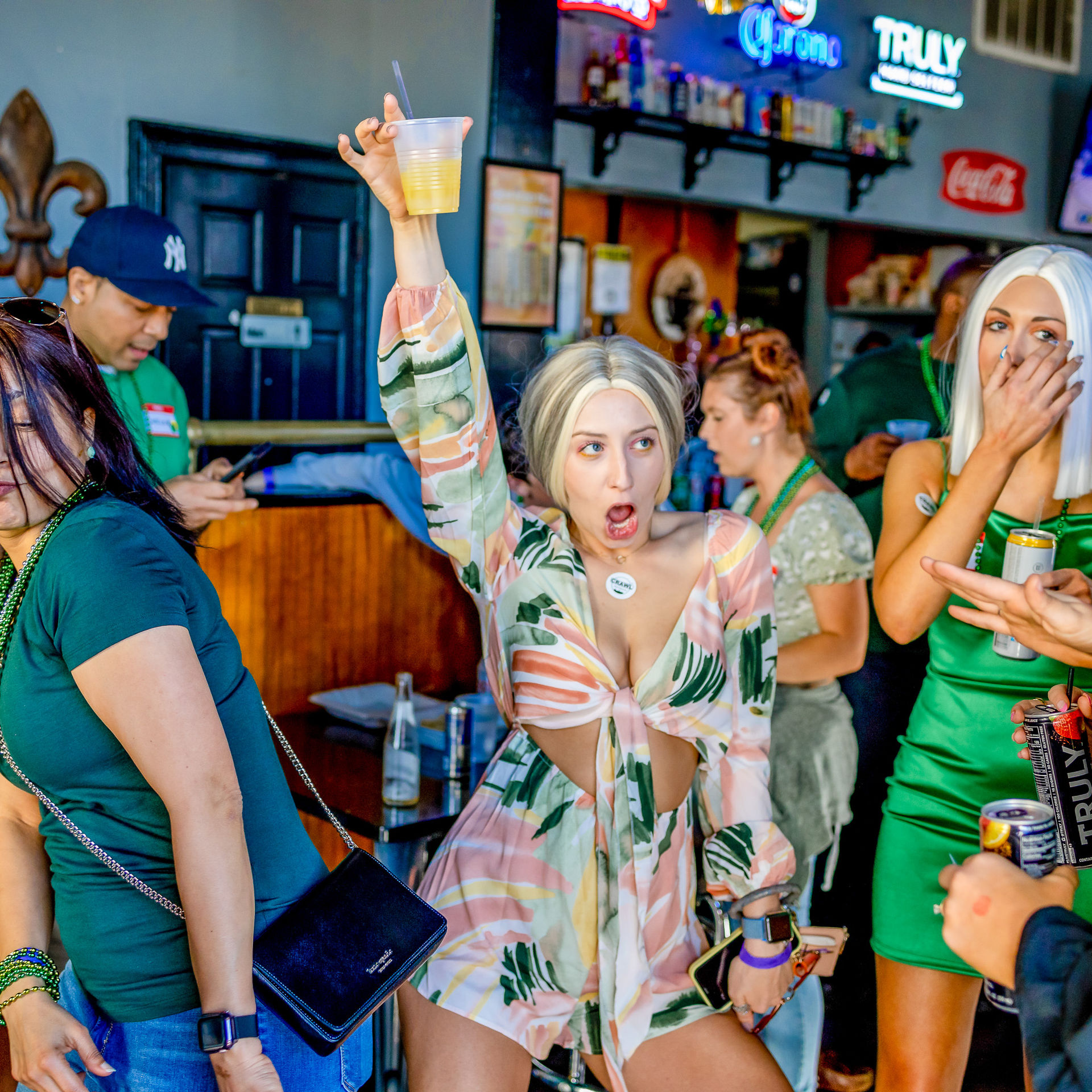 Excited woman in a tropical tie-front dress raises a plastic cup while cheering at a crowded bar St. Patrick’s Day celebration, surrounded by people in green and party beads in a lively pub interior