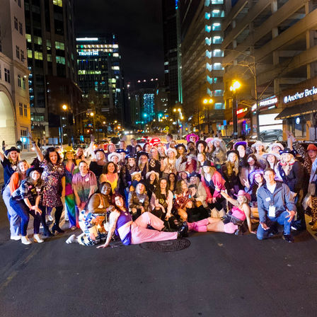 Large group in colorful costumes and hats posing across a downtown street at night with city lights and skyscrapers glowing behind them, lively urban celebration.