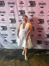 Smiling woman in a short white halter dress, veil, and sparkly sandals posing on a concrete floor in front of a branded step-and-repeat backdrop at a New Orleans nightlife event.