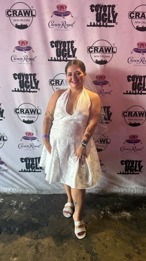 Smiling woman in a short white halter dress, veil, and sparkly sandals posing on a concrete floor in front of a branded step-and-repeat backdrop at a New Orleans nightlife event.