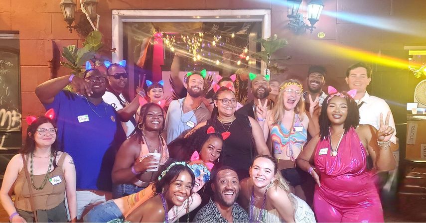 Group of friends in colorful outfits and glowing cat ears posing and laughing at a lively outdoor patio party under string lights