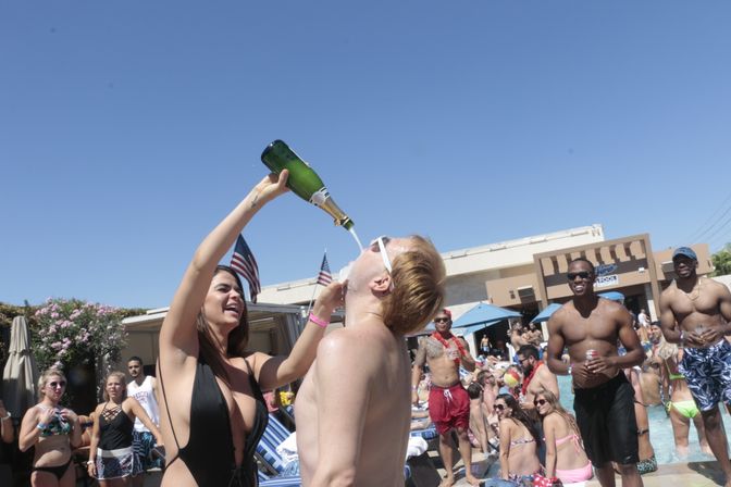 Outdoor pool party at a resort on a sunny day — woman pouring champagne into a man’s mouth while a crowd in swimsuits cheers