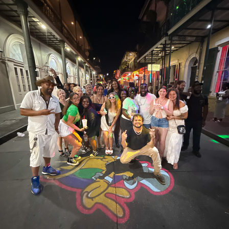 Large group of friends posing on a colorful painted street mural under neon lights and wrought-iron balconies in the New Orleans French Quarter at night