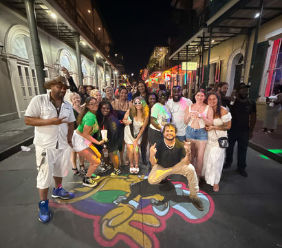 Large group of friends posing on a colorful painted street mural under neon lights and wrought-iron balconies in the New Orleans French Quarter at night