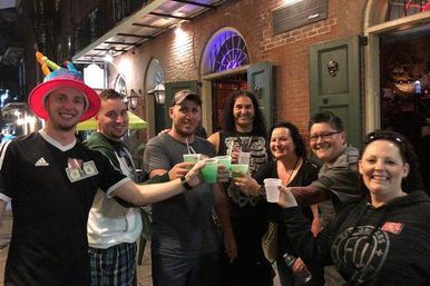 Group of seven friends smiling and clinking bright green frozen drinks in plastic cups outside a brick bar at night, lively nightlife scene