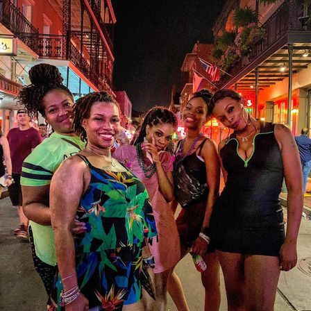 Group of five women posing on a lively Bourbon Street night in New Orleans, smiling with beads, neon lights and wrought-iron balconies in the background