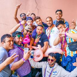 Group of friends at a colorful outdoor party wearing Mardi Gras beads, sunglasses and name tags, posing with an inflatable flamingo and drinks against a pink wall.