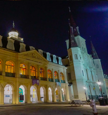 Nighttime scene in New Orleans' French Quarter: illuminated colonial-style building with arched arcades and glowing windows beside a tall church with twin spires, empty benches in the plaza.