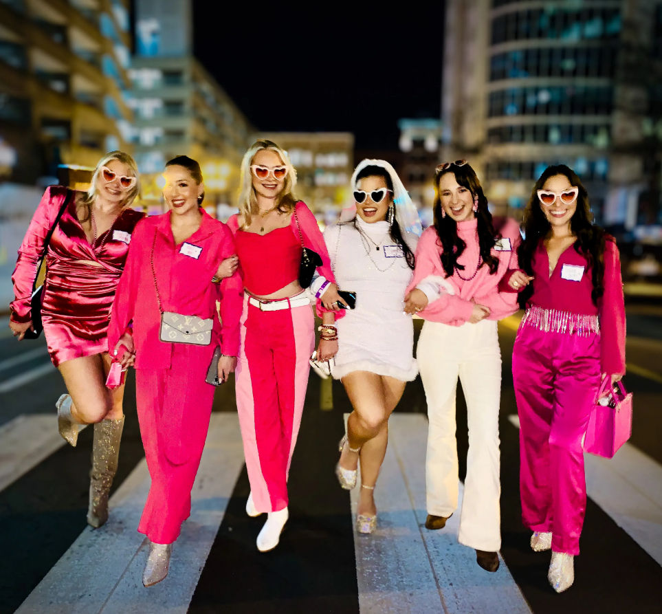 Six friends walking arm-in-arm across a city crosswalk at night, wearing coordinated pink outfits and heart-shaped sunglasses, one in white with a veil, smiling and celebrating a night out.