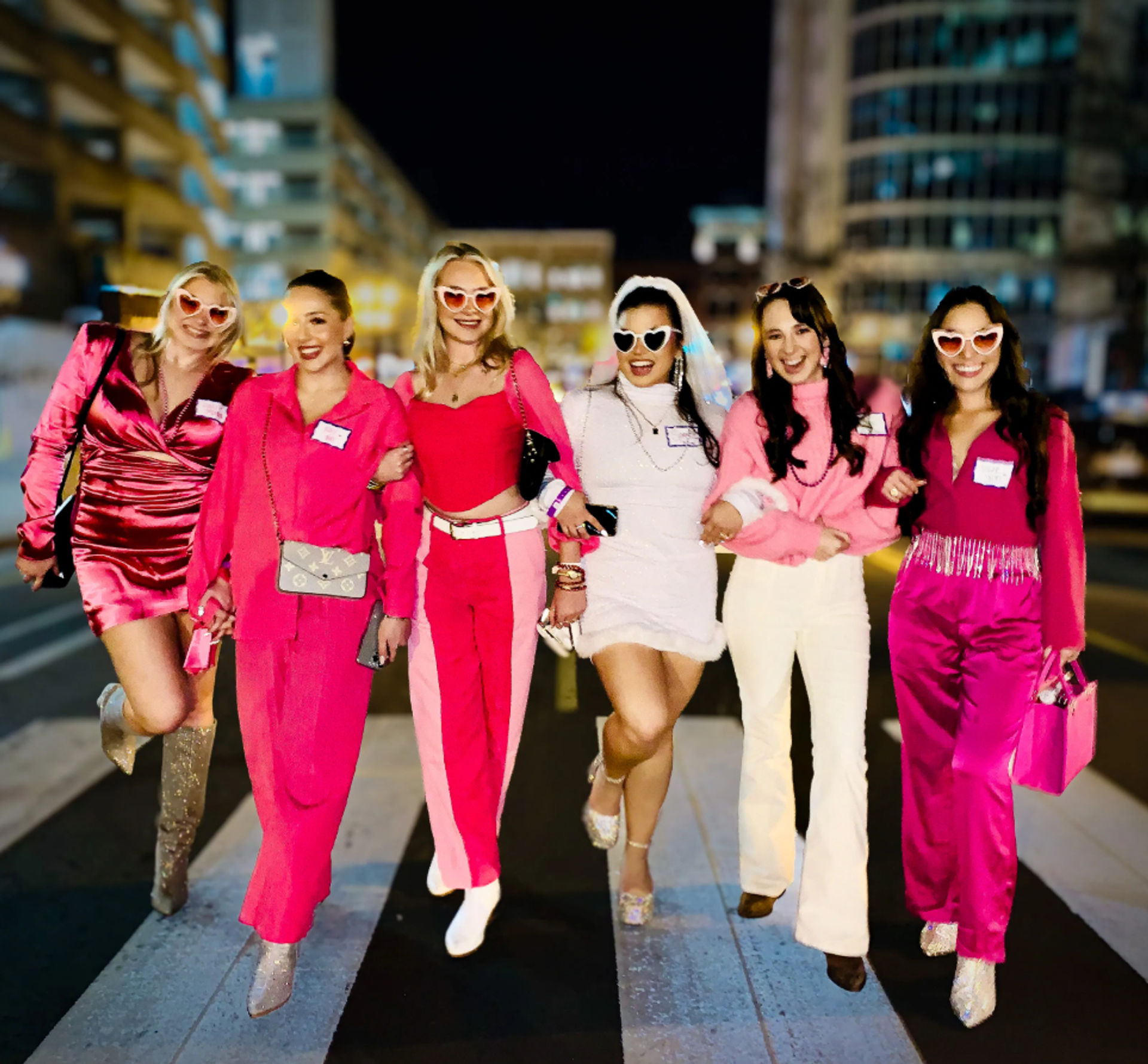 Six friends walking arm-in-arm across a city crosswalk at night, wearing coordinated pink outfits and heart-shaped sunglasses, one in white with a veil, smiling and celebrating a night out.
