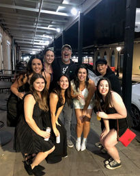 Nine friends posing on a covered city sidewalk at night, smiling and holding drinks; bride-to-be in a white dress, veil and sparkly boots surrounded by friends in black party outfits under overhead lights — urban nightlife scene.