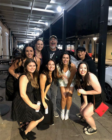 Nine friends posing on a covered city sidewalk at night, smiling and holding drinks; bride-to-be in a white dress, veil and sparkly boots surrounded by friends in black party outfits under overhead lights — urban nightlife scene.