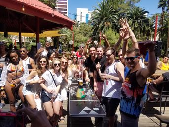 Sunny outdoor pool party with a crowd cheering at a resort-style bar, palm trees, string lights and city high-rises in the background.