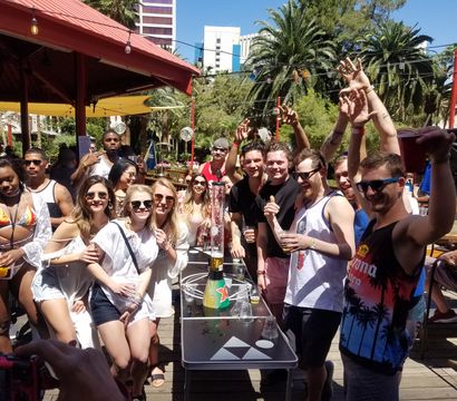 Sunny outdoor pool party with a crowd cheering at a resort-style bar, palm trees, string lights and city high-rises in the background.