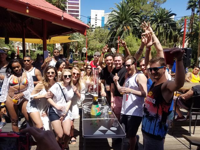 Sunny outdoor pool party with a crowd cheering at a resort-style bar, palm trees, string lights and city high-rises in the background.