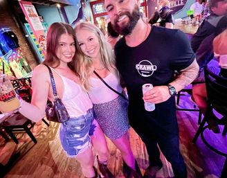 Three friends smiling and holding drinks in a colorful neon-lit bar, two women in summer outfits and a bearded man in a black shirt.
