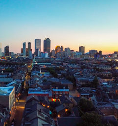 Aerial view of New Orleans skyline at sunset, glowing downtown skyscrapers rising behind historic low-rise French Quarter rooftops and lit streets.