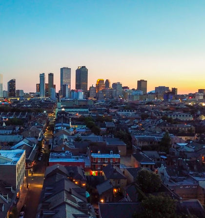 Aerial view of New Orleans skyline at sunset, glowing downtown skyscrapers rising behind historic low-rise French Quarter rooftops and lit streets.