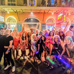 Large group of people in colorful wigs and party outfits smiling and posing on a neon-lit downtown street at night under a balcony — lively city nightlife scene.
