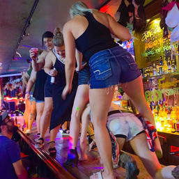 Women dancing on a neon-lit bar top in a crowded nightlife scene, wearing shorts and dresses and holding drinks amid colorful lights and bottles.