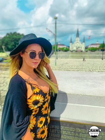 Sunflower-dress traveler in a black hat and round sunglasses posing by Jackson Square with St. Louis Cathedral spires and a cloudy blue sky in New Orleans