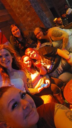 Smiling group selfie of friends at a cozy indoor bar — warm orange lighting, exposed brick wall, cocktails and visible tattoos for a lively night-out vibe.