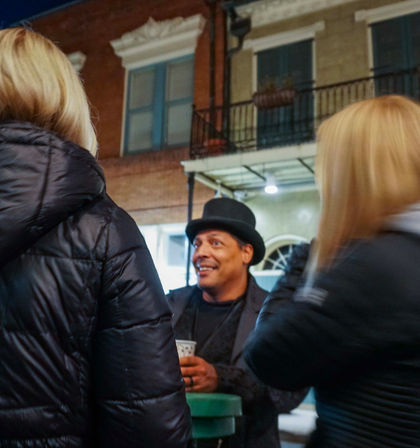 Smiling man in a bowler hat holding a cup chats with two women in puffy jackets on a city sidewalk at night in front of a brick building with a balcony