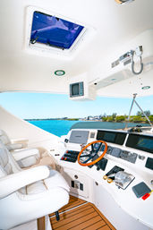 Yacht helm with white leather captain seats, wooden steering wheel, navigation displays and throttle controls overlooking turquoise water and a tree-lined marina coastline