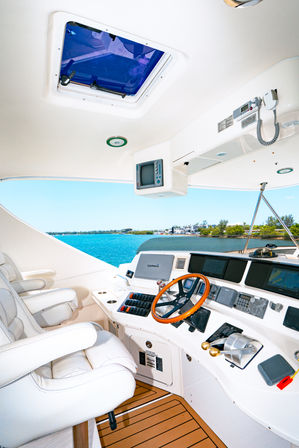 Yacht helm with white leather captain seats, wooden steering wheel, navigation displays and throttle controls overlooking turquoise water and a tree-lined marina coastline