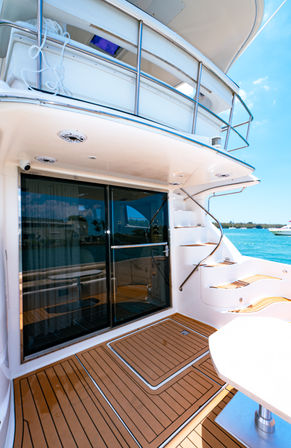 Luxury yacht aft deck with teak flooring, sliding glass doors and stainless spiral stairs overlooking turquoise marina waters under a sunny sky.