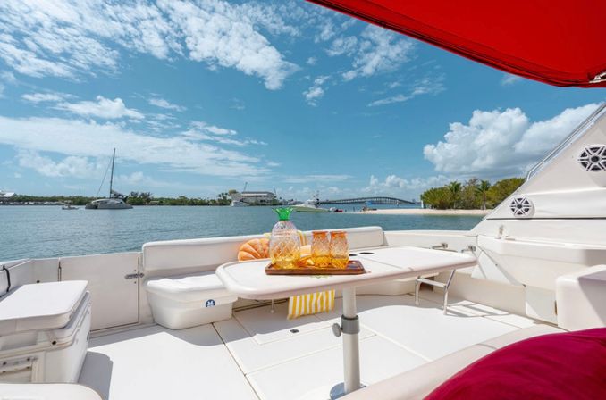 Yacht deck with pineapple pitcher and amber glasses under a red sunshade, overlooking sailboats, a bridge and palm-lined coastal inlet on a sunny day.