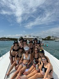 Bachelorette boat party on a luxury yacht at a sunny coastal marina — group of friends in swimsuits and matching bucket hats, bride wearing a sparkling 'BRIDE' cap and holding drinks under a blue sky.