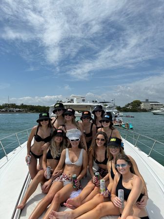Bachelorette boat party on a luxury yacht at a sunny coastal marina — group of friends in swimsuits and matching bucket hats, bride wearing a sparkling 'BRIDE' cap and holding drinks under a blue sky.