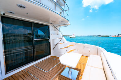 Aft deck of a luxury yacht with teak flooring, cushioned bench and small white table, curved stairs to the flybridge, sliding glass doors, and bright turquoise sea under a sunny sky.