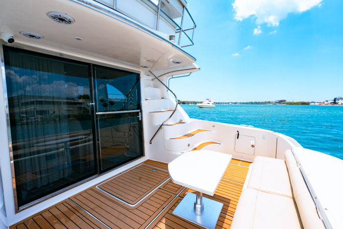 Aft deck of a luxury yacht with teak flooring, cushioned bench and small white table, curved stairs to the flybridge, sliding glass doors, and bright turquoise sea under a sunny sky.