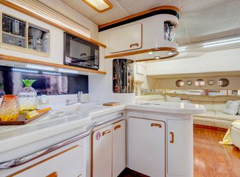 Bright luxury yacht galley with white curved cabinetry and wood trim, microwave, sink, decorative glassware, LED under-cabinet lighting, and open plan to a cream leather salon with porthole windows and wood flooring