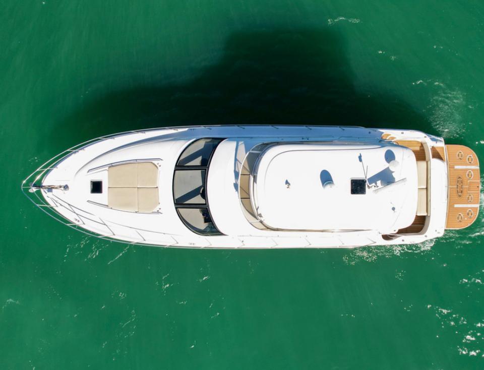 Aerial top-down drone view of a sleek white luxury yacht gliding on emerald green water, showing teak swim platform, foredeck sun pad, and curved windshield