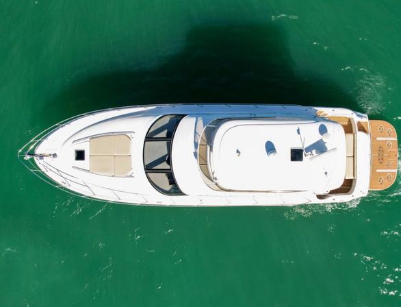 Aerial top-down drone view of a sleek white luxury yacht gliding on emerald green water, showing teak swim platform, foredeck sun pad, and curved windshield