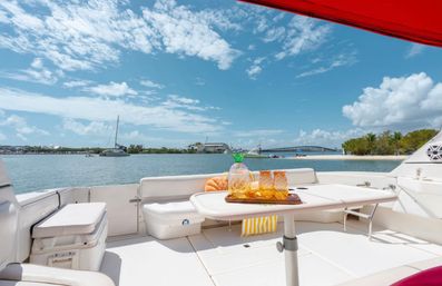 Sunny yacht deck with white seating and a table holding a pineapple-shaped pitcher and amber glasses on a tray, overlooking a calm turquoise harbor with a sailboat, palm-lined shoreline and a low bridge under a bright blue sky.