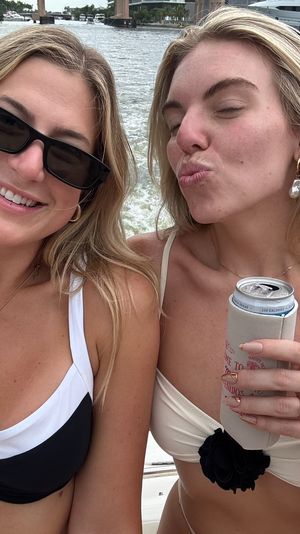 Two women in bikinis smiling and posing on a boat cruise, one holding a canned drink, marina docks and yachts in the background.