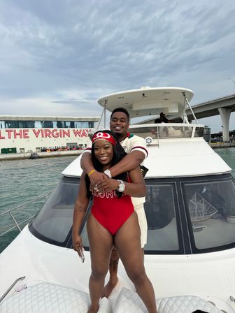 Smiling couple hugging on the bow of a white yacht in a marina, waterfront terminal and highway bridge visible under a cloudy sky