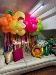 Indoor bridal shower party decor in a cozy living room: vibrant neon pink, orange, yellow and green balloon garland with paper rosettes, multicolored streamers, a "Bride-to-be" neon sign, gold and magenta pillows on a cream sofa and novelty inflatable balloons.