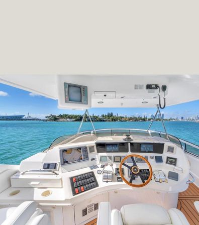 Sunny yacht helm with wooden steering wheel, modern navigation screens and controls on a white dashboard, teak deck, turquoise bay water and distant coastal skyline under a clear blue sky.