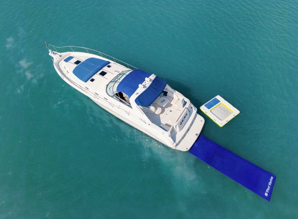 Aerial view of a white luxury yacht with blue canopy anchored in clear turquoise water, a long blue swim mat trailing from the stern and a yellow-edged floating trampoline nearby.