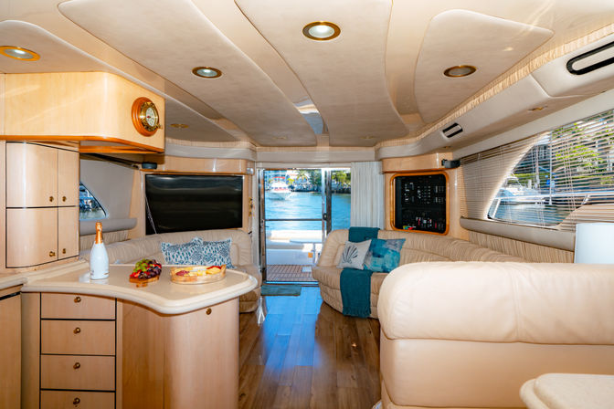 Sunlit luxury yacht salon with cream leather sofas, wood flooring, curved light-wood cabinetry, counter with champagne and snacks, blue throw pillows and sliding doors opening to a marina water view